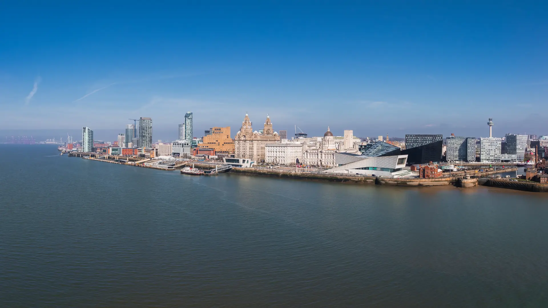 Aerial view of Liverpool, England, highlighting the Three Graces, the Museum of Liverpool, modern architecture, and the River Mersey under a clear sky.