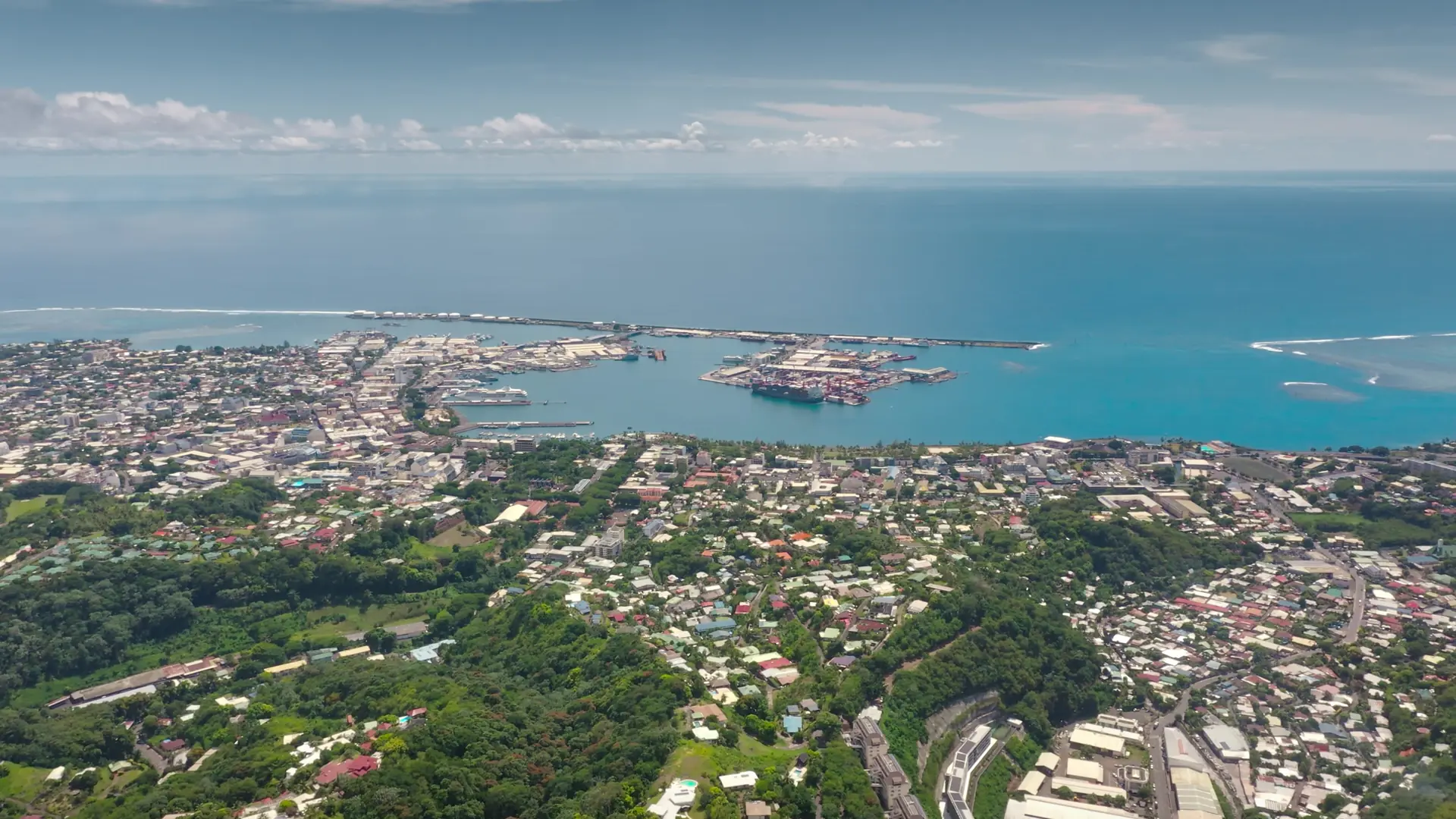 Aerial drone fly over Papeete coast, Tahiti cityscape harbor, green mountain hills, colorful village houses, streets, buildings, lush tropical vegetation against pacific ocean backdrop. Remote travel