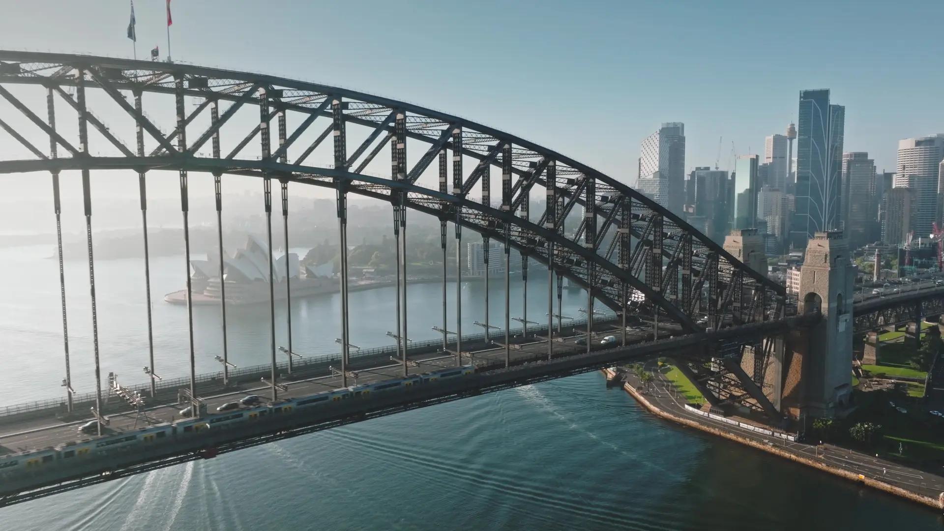 Australia, Sydney: Aerial view of Sydney Harbour Bridge, Sydney Opera House and city skyline in background. Sunny morning light, train and cars crossing the bridge. Modern architecture. Drone